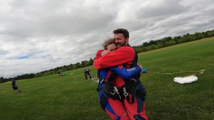 Demande en mariage chez Parachute Voltige Un homme et une femme habillés de harnais de parachute tandem et de combinaisons de saut rouge s'enlacent après une demande en mariage au moment de l'atterrissage.