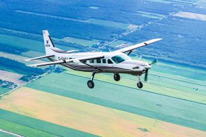 Un Cessna Super Caravan prend sont envol au dessus de champs de toutes les teintes de verts pour aller larger des parachutistes au dessus de Parachute Voltige.